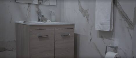 Une salle de bain avec un mur en marbre, un meuble-lavabo en bois doté de deux tiroirs, un miroir et un porte-rouleau de papier toilette.