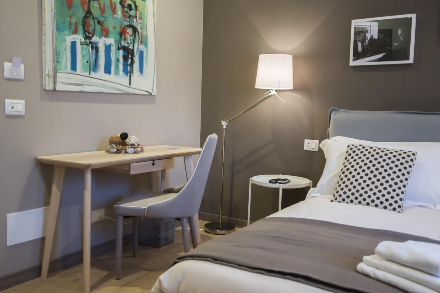 A hotel room with a bed, a desk, a chair, a lamp, and a painting on the wall.