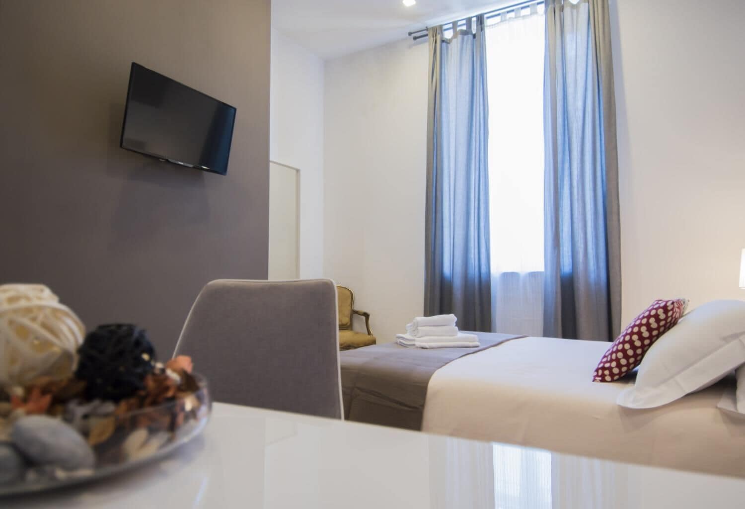 Une chambre d’hôtel avec un lit, un téléviseur fixé au mur, une table à manger avec une assiette de fruits et une fenêtre avec des rideaux.