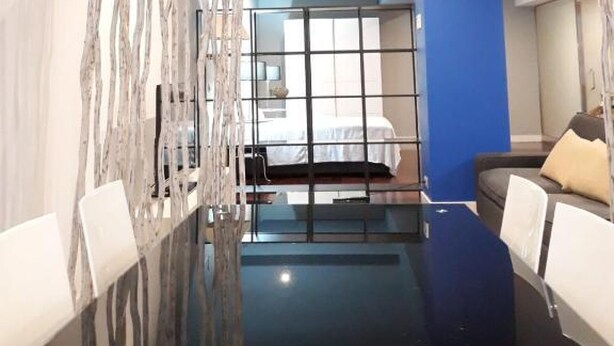A modern hotel room with a glass partition, a bed, a blue accent wall, and a seating area with a sofa and chairs.