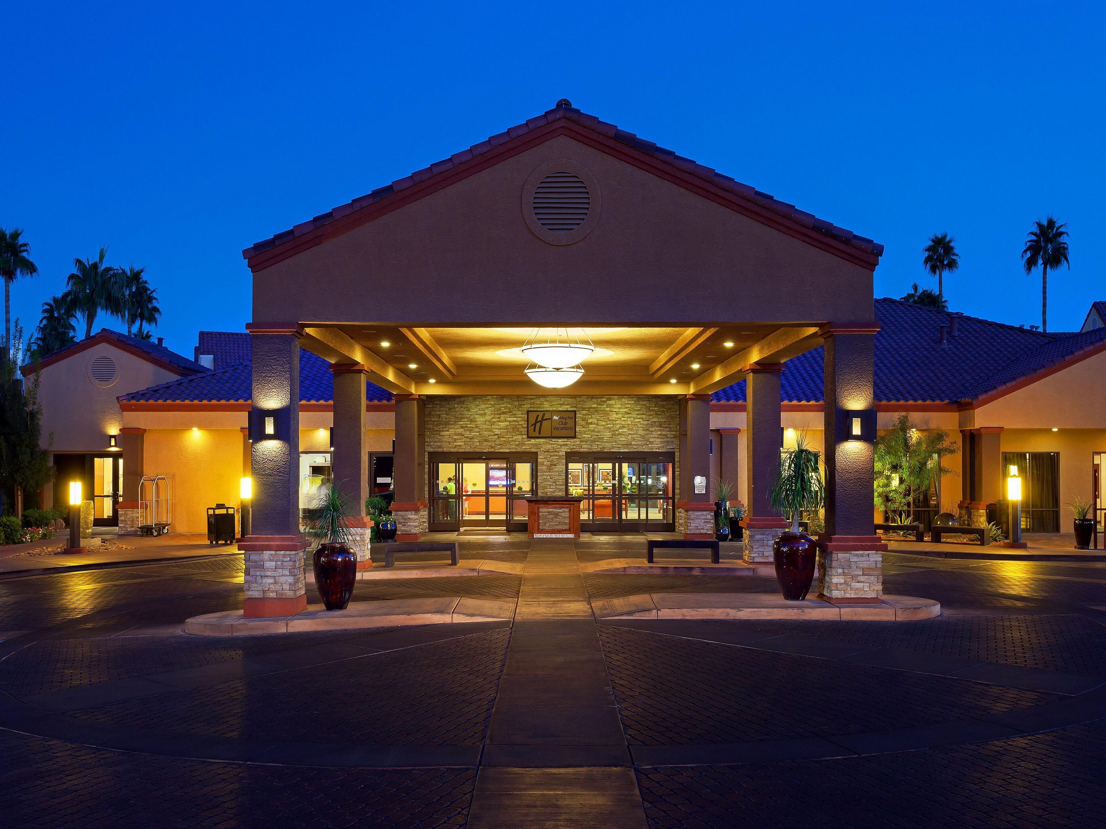 A hotel entrance with a covered walkway, palm trees, and a well-lit reception area.