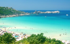 Una playa con aguas turquesas cristalinas, afloramientos rocosos y una multitud de personas disfrutando del sol y el mar.
