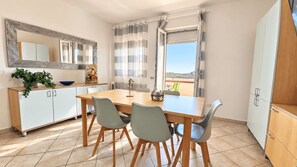 A dining area with a wooden table and four chairs, a white cabinet with a plant, a mirror, and a window with curtains. - Case Vacanze Santa Teresa Gallura (Santa Teresa di Gallura)