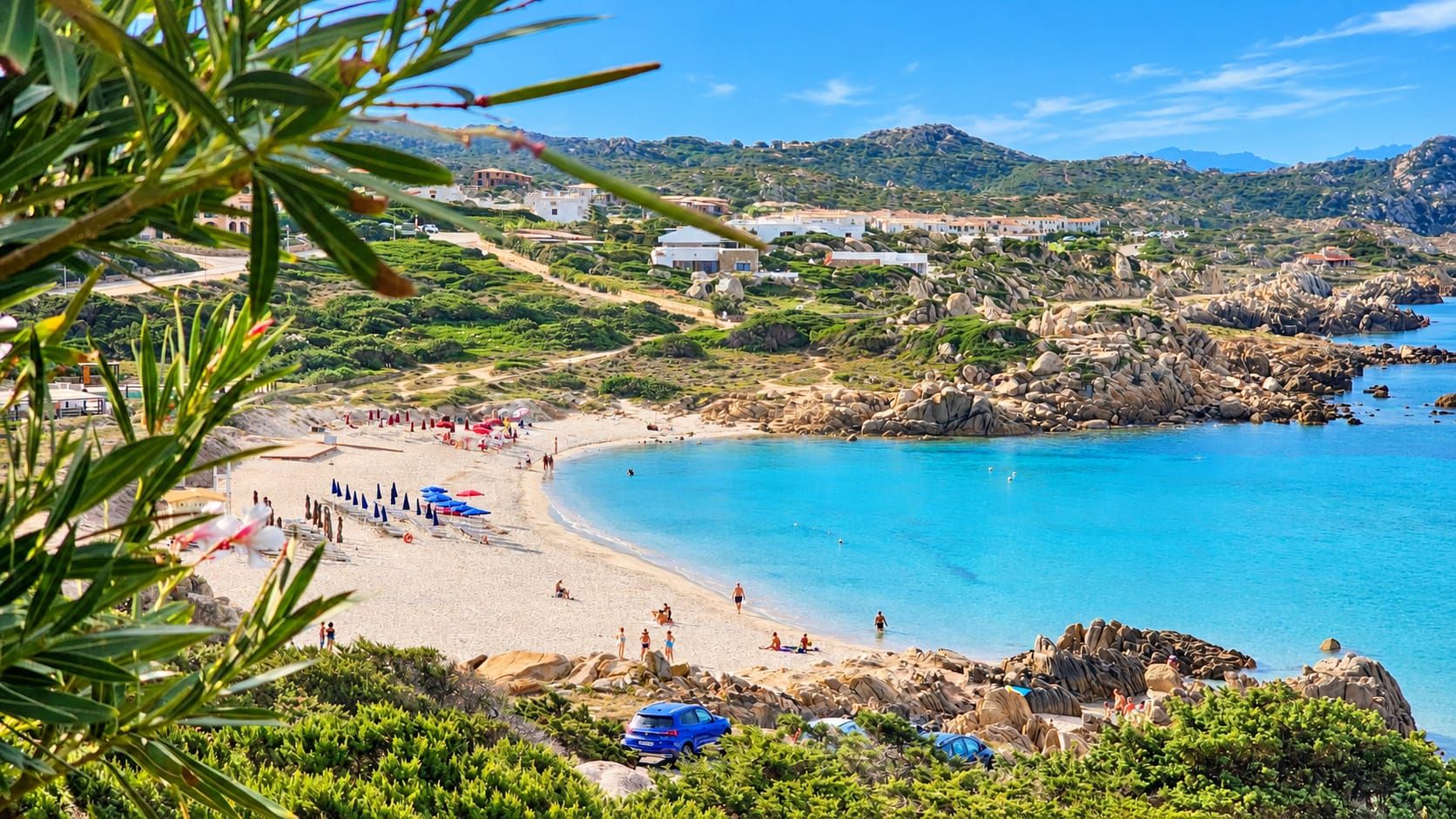 Una playa costera con aguas azul turquesa, terreno rocoso y edificios a lo lejos.