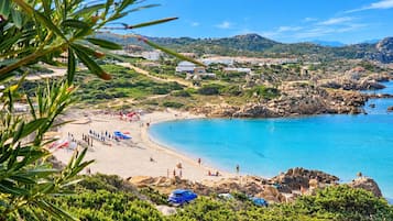 Uma praia costeira com águas azuis cristalinas, terreno rochoso e vista de edifícios ao longe.