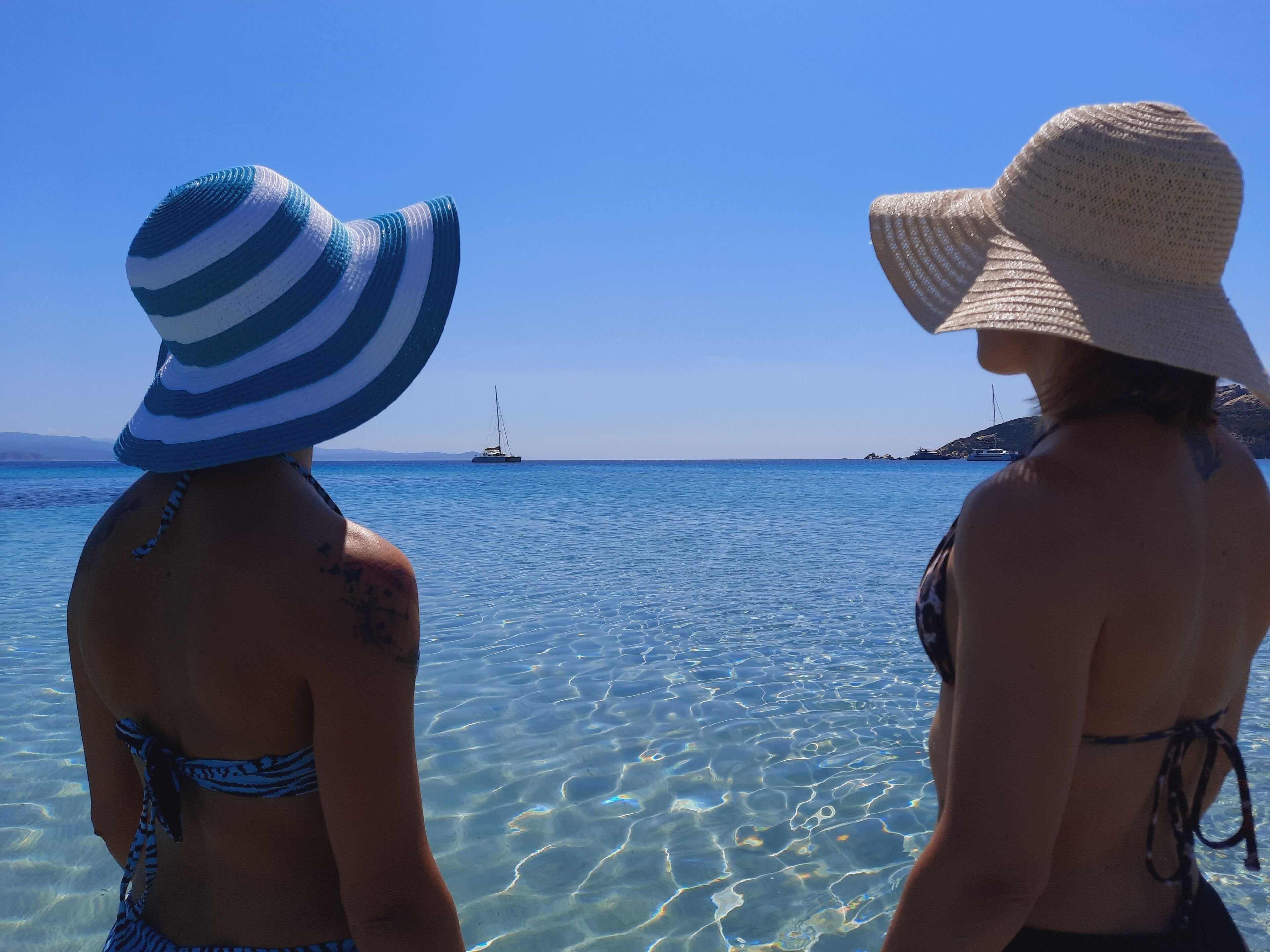 Two people in swimsuits and hats by a clear sea with a sailboat in the distance.