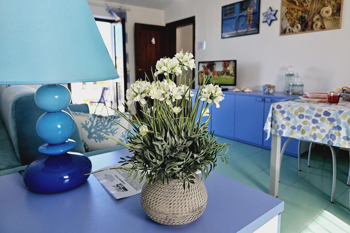 Ein Zimmer mit einem blauen Sofa, einem Tisch mit einer Lampe, einer Vase mit weißen Blumen und einer Küchenzeile im Hintergrund.