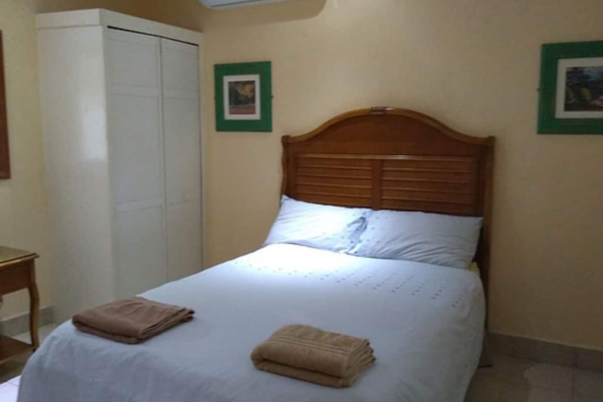 A neatly made bed with a wooden headboard, a white wardrobe, and two framed pictures on the wall.