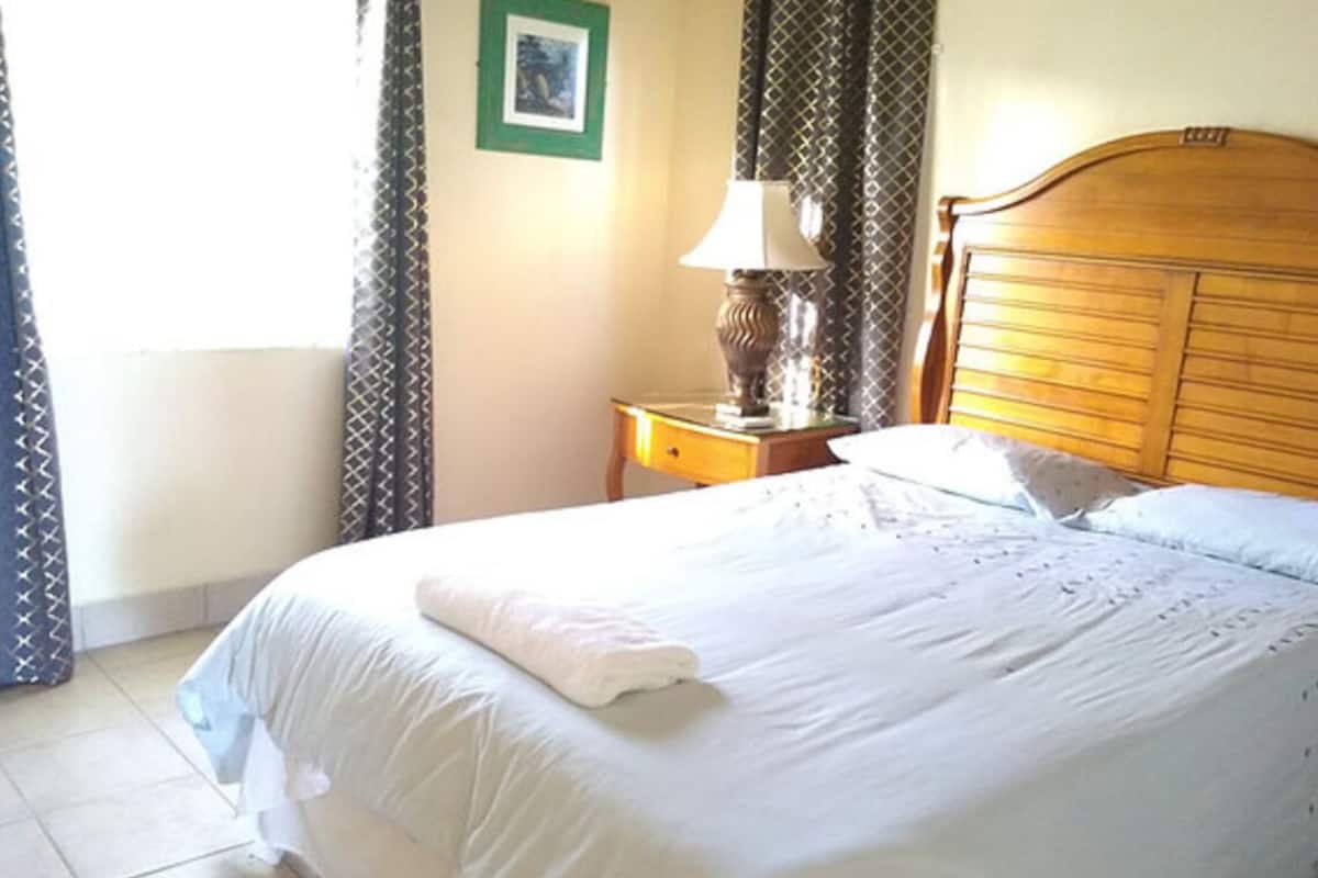 A neatly made bed with a white comforter and a folded white towel. There is a wooden nightstand with a lamp and a framed picture on the wall.