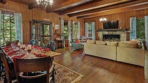 A wooden interior with a dining area, sofa, fireplace, and large windows.