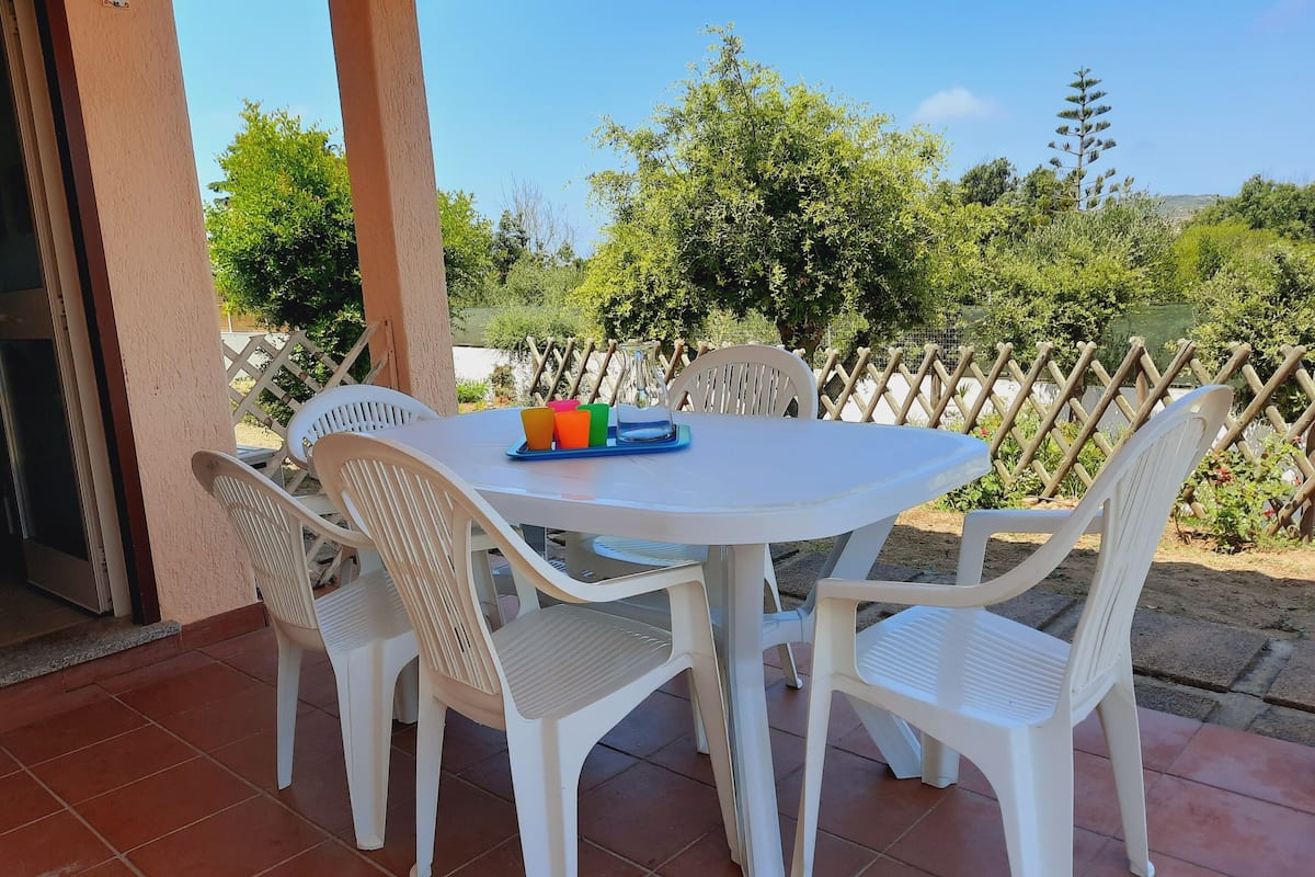 Un patio con una mesa redonda y cuatro sillas, una bandeja con tazas de colores, y vistas a follaje y árboles.