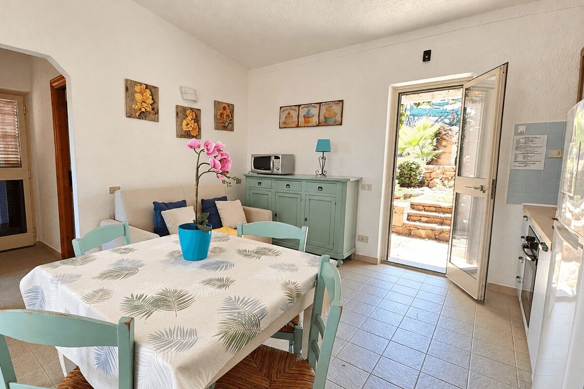 A dining area with a table set for two, a blue vase with flowers, and a kitchen area with a microwave and cabinets.