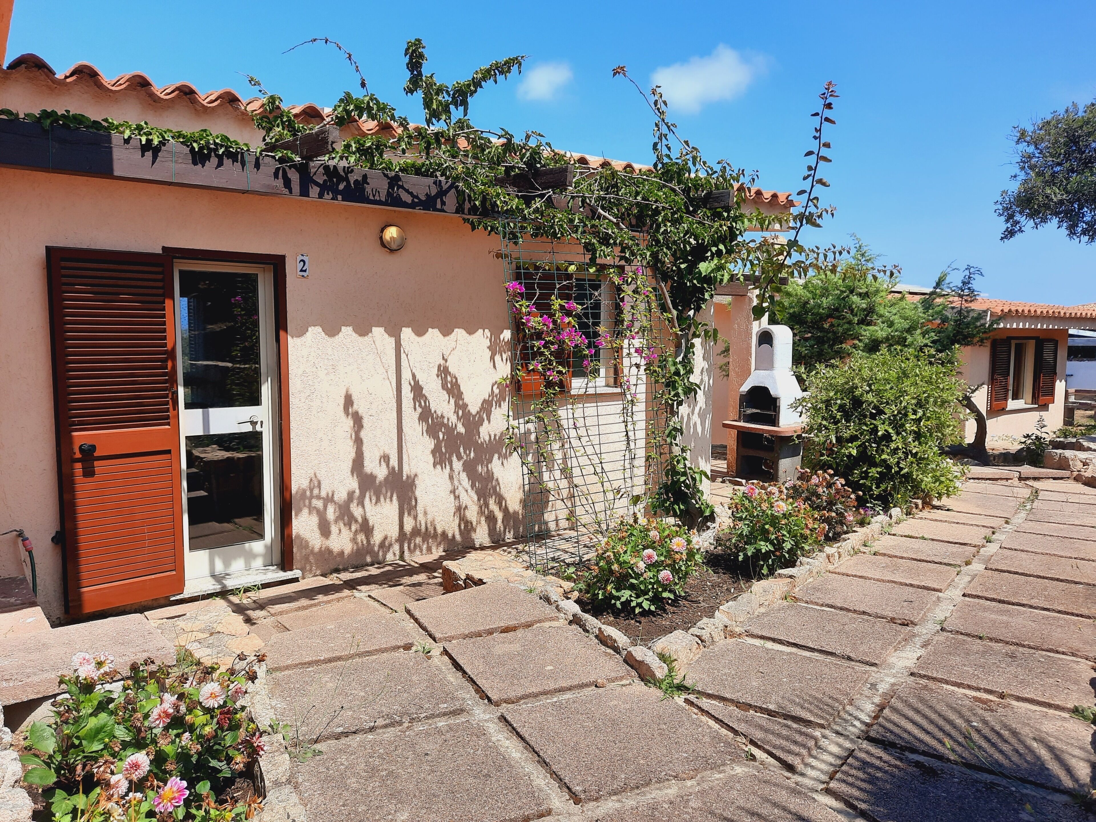 A house with a tiled roof, a door with a window, a small garden with flowers, and a barbecue grill.
