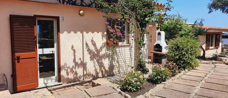 A house with a tiled roof, a door with a window, a small garden with flowers, and a barbecue grill.
