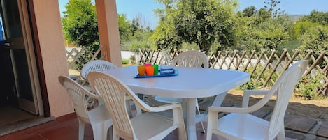 Eine Terrasse mit weißem Tisch und Stühlen, einer Glastür und Blick auf Grünflächen und einen Zaun.