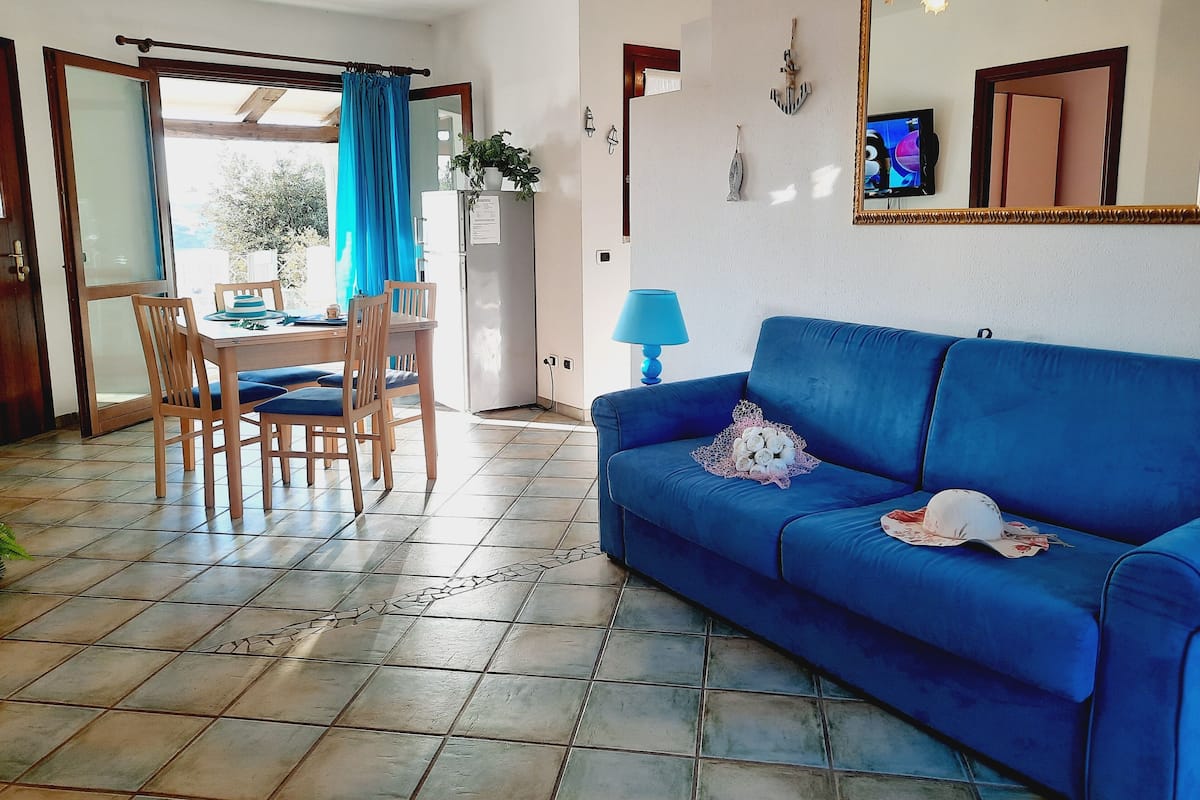 Una sala de estar con un sofá azul, una mesa de comedor con sillas, una lámpara azul y un espejo grande en la pared.