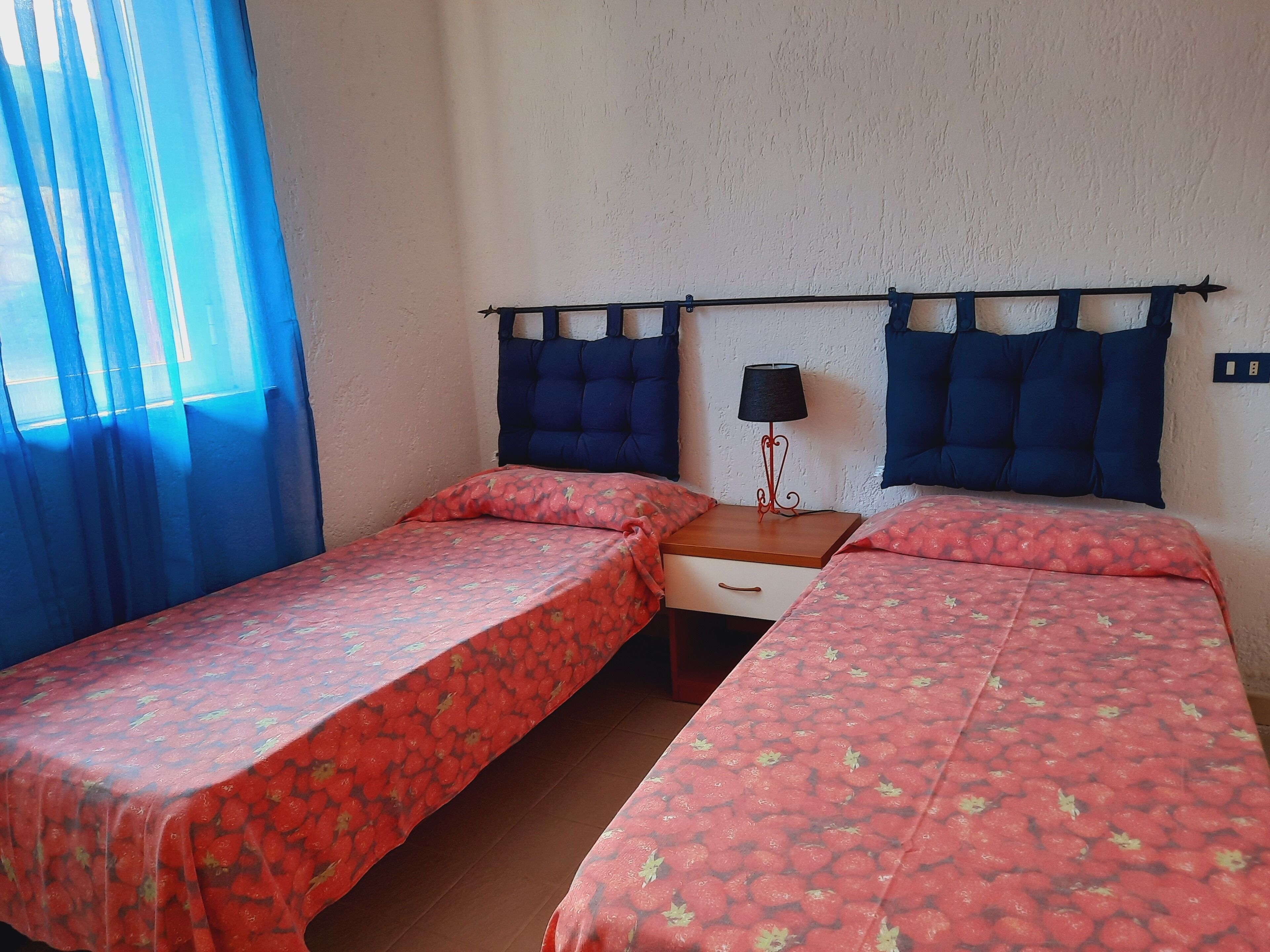 A room with two single beds, a nightstand with a lamp, and a window with blue curtains.