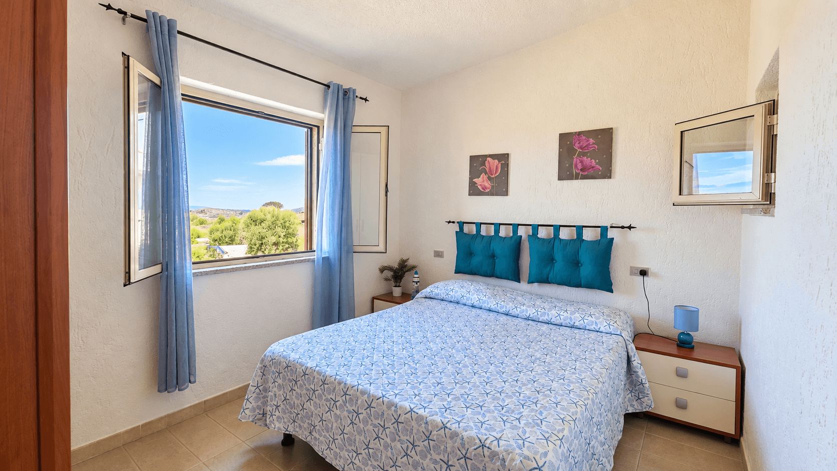 Een slaapkamer met een bed, gordijnen, een nachtkastje, een lamp en kunst aan de muur.