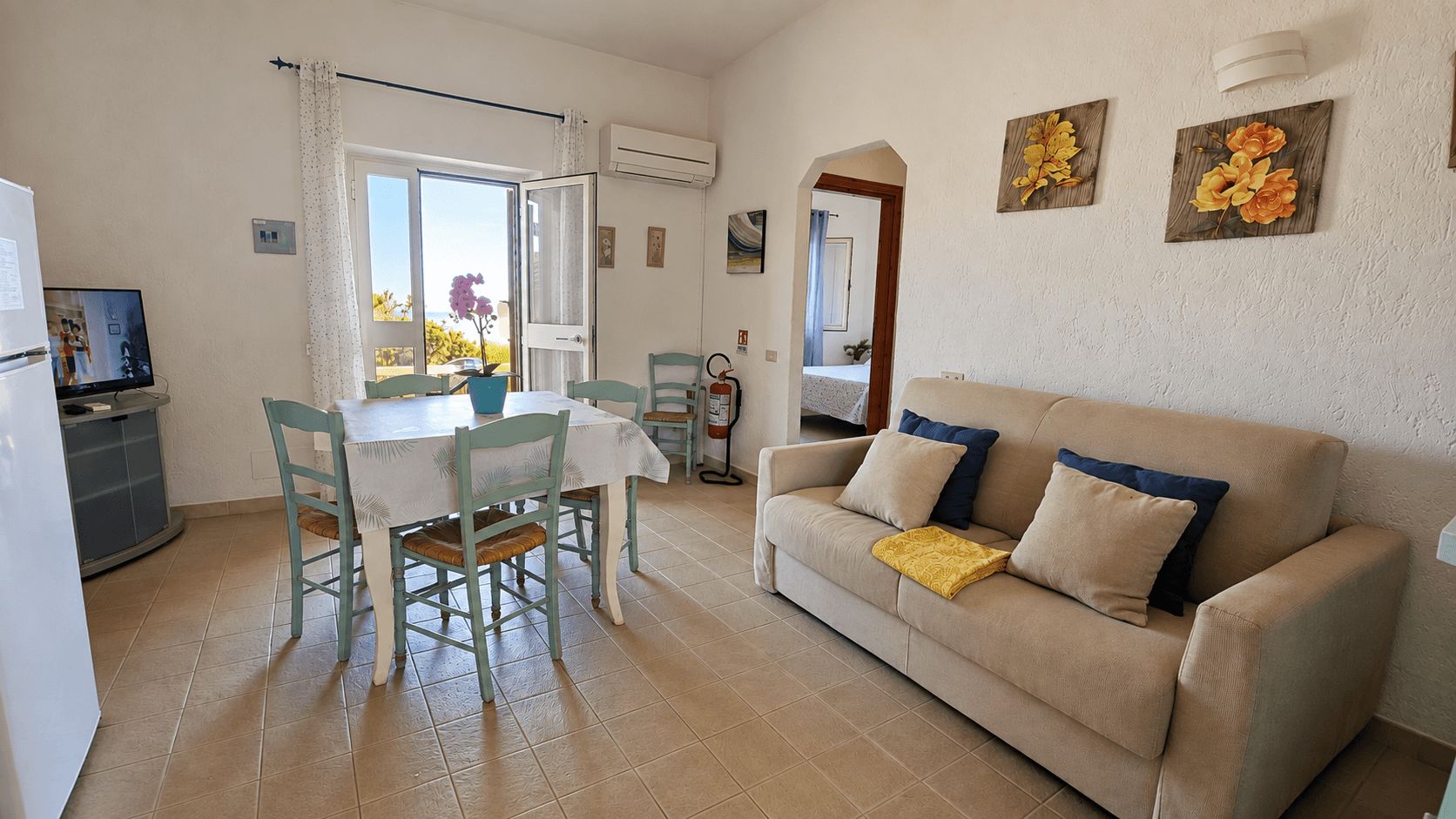 A living room with a beige sofa, a dining table with chairs, and a kitchen area with a refrigerator.