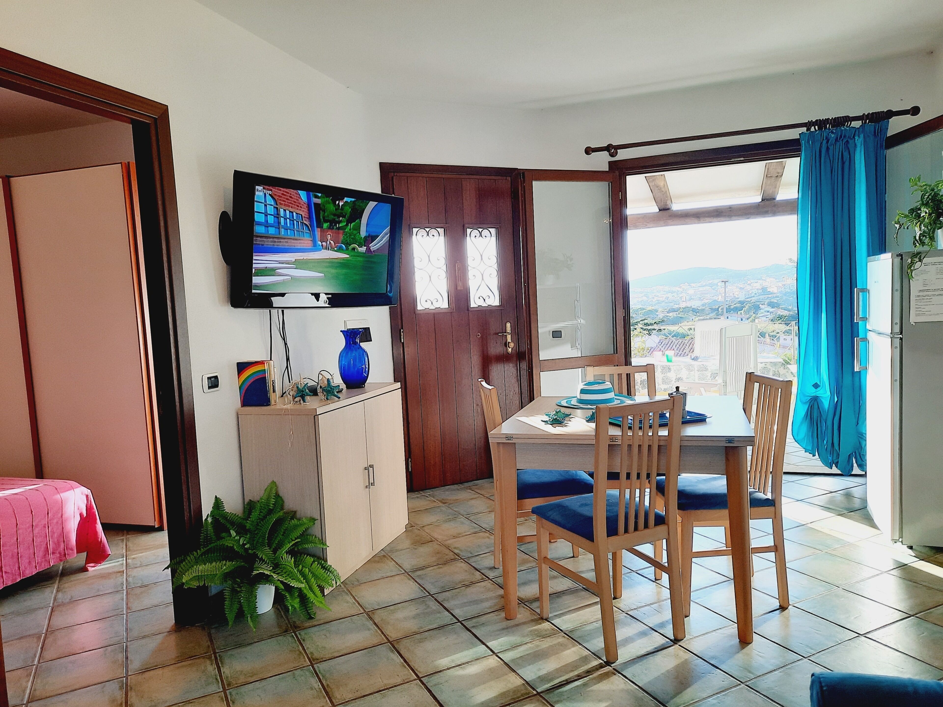 A dining area with a table and chairs, a television mounted above, a cabinet with a vase, and a view through a door.