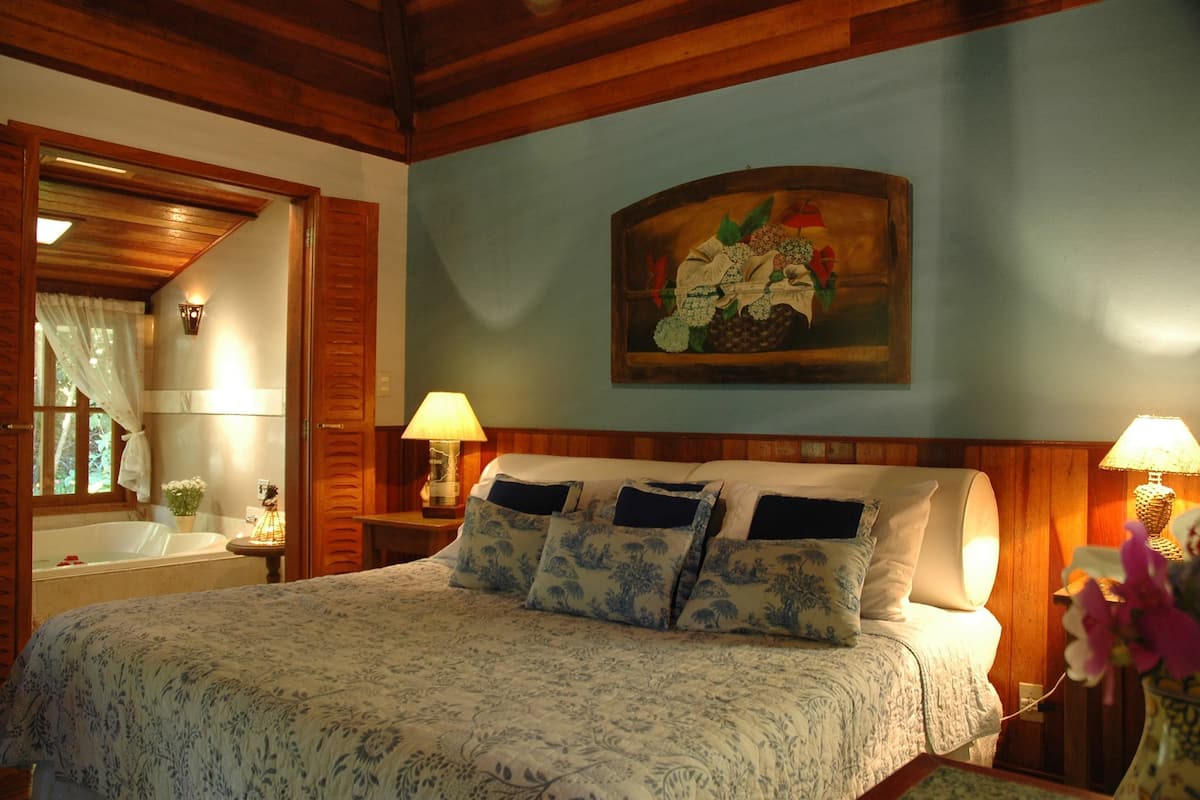 Quarto com uma cama grande, cabeceira de madeira, duas luminárias de cabeceira, um quadro na parede e uma janela com cortinas.