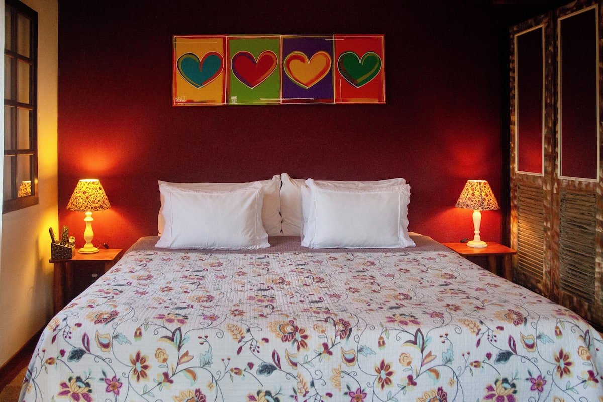 Quarto de hotel com uma cama grande, colcha florida, duas luminárias de cabeceira e uma parede com decorações em formato de coração.