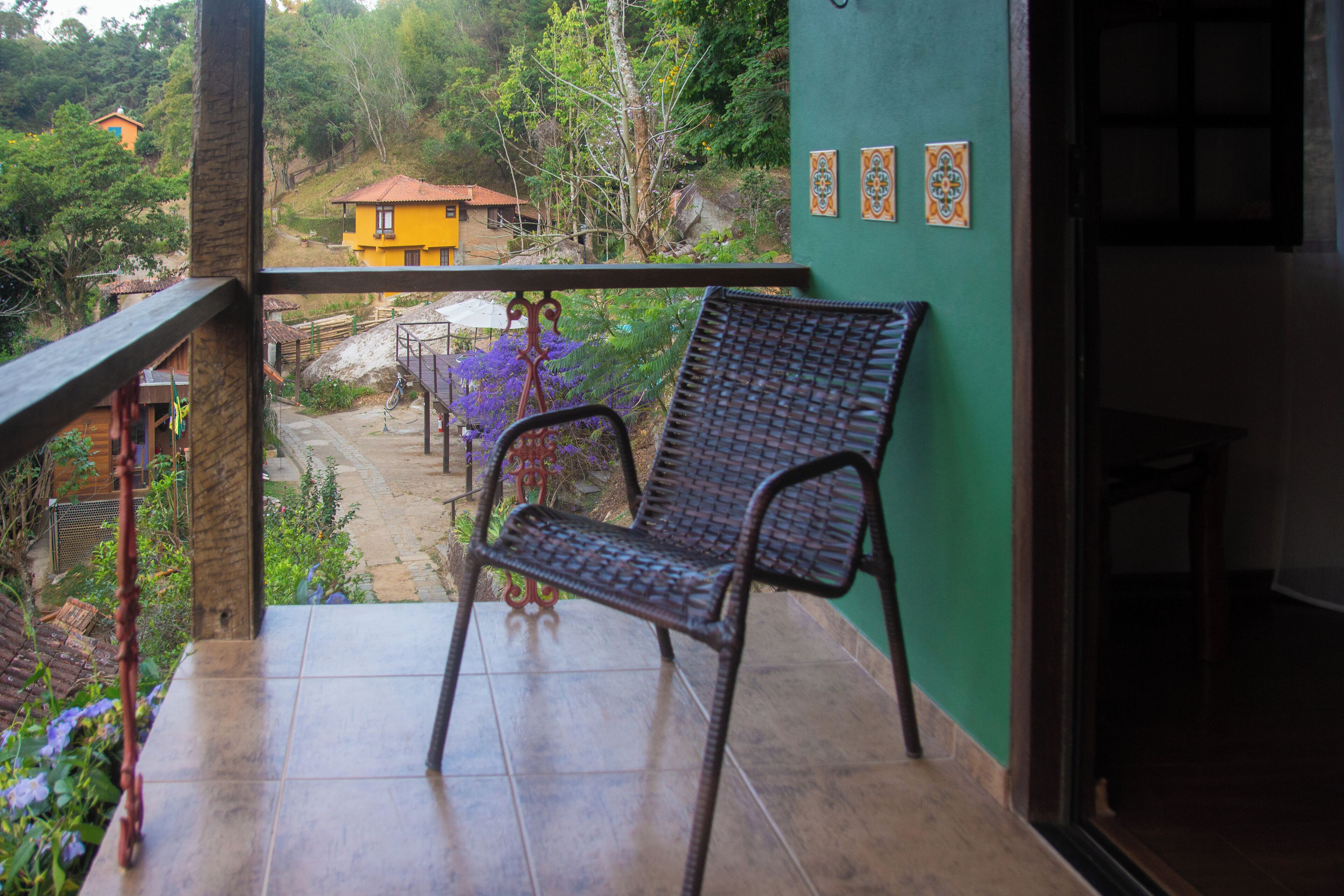Uma varanda com cadeira de vime, vista de um edifício amarelo e parede verde com azulejos decorativos.