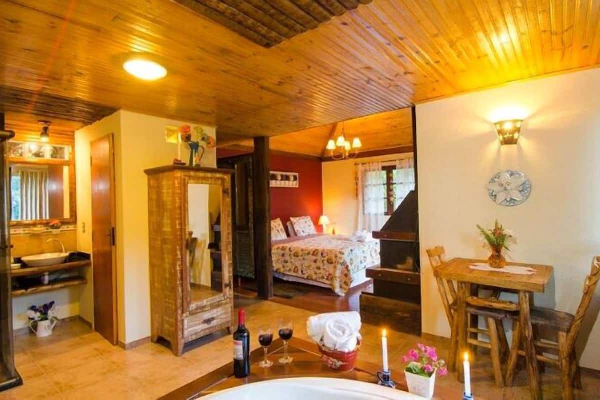 Quarto aconchegante com teto de madeira, uma cama com roupa de cama florida, uma mesa de madeira com um vaso e um relógio de parede.