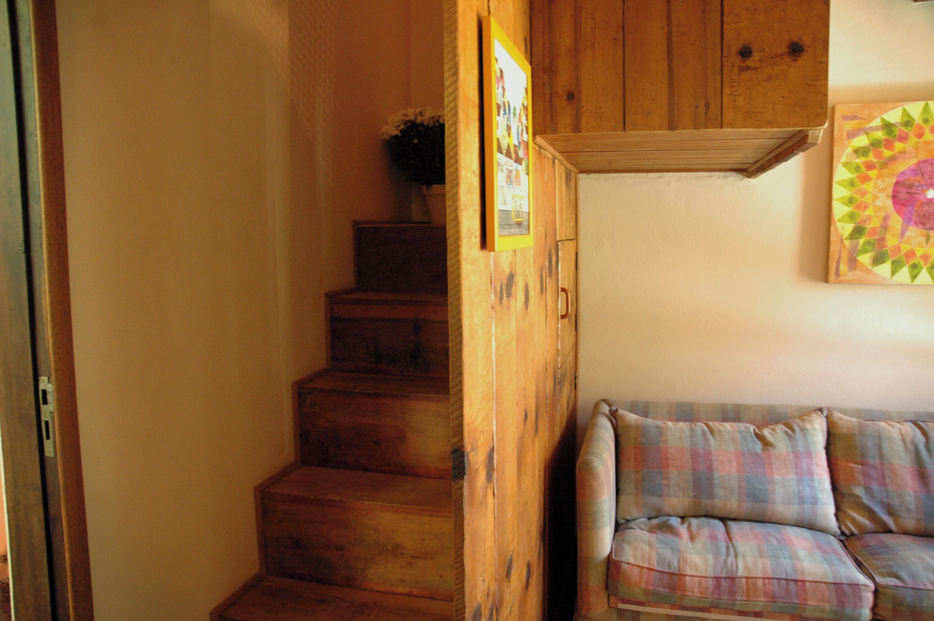 Una escalera de madera que baja a un nivel inferior, un armario de madera y un sofá de cuadros en una habitación con una pared lisa y un cuadro enmarcado.