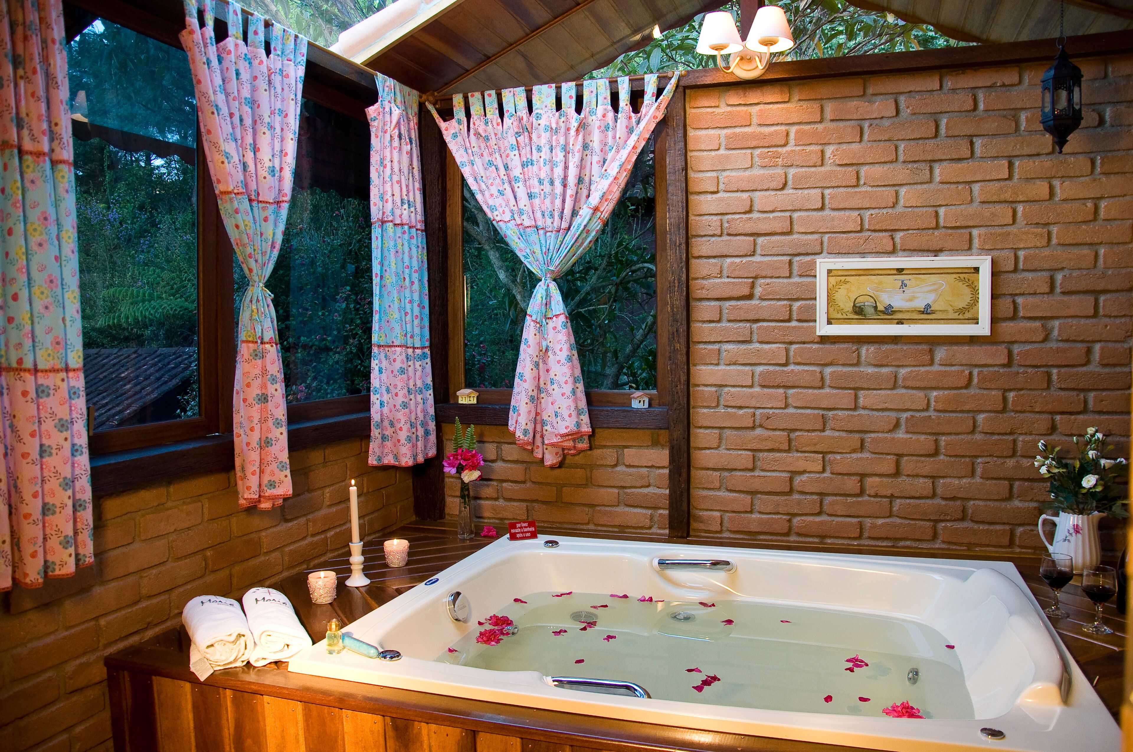 A bathroom with a large bathtub, floral curtains, a brick wall, and a framed picture.