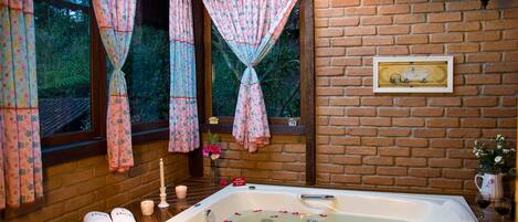 A bathroom with a large bathtub, floral curtains, a brick wall, and a framed picture.