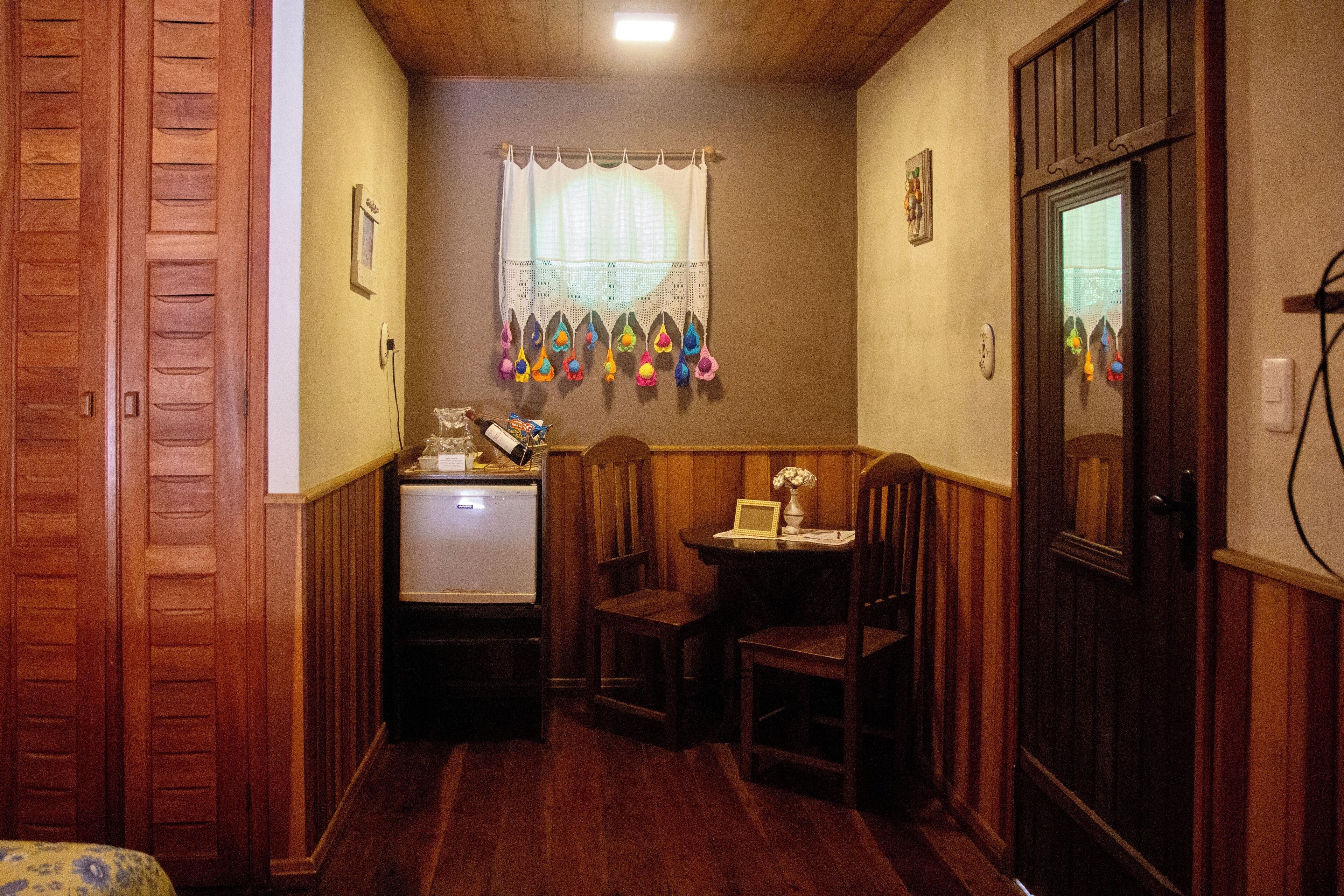 A narrow hallway with wooden walls and a wooden door on the right. There is a small table with a lamp and a picture frame on it. A window with a curtain is visible in the background.