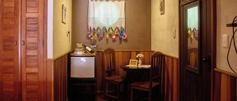 A narrow hallway with wooden walls and a wooden door on the right. There is a small table with a lamp and a picture frame on it. A window with a curtain is visible in the background.