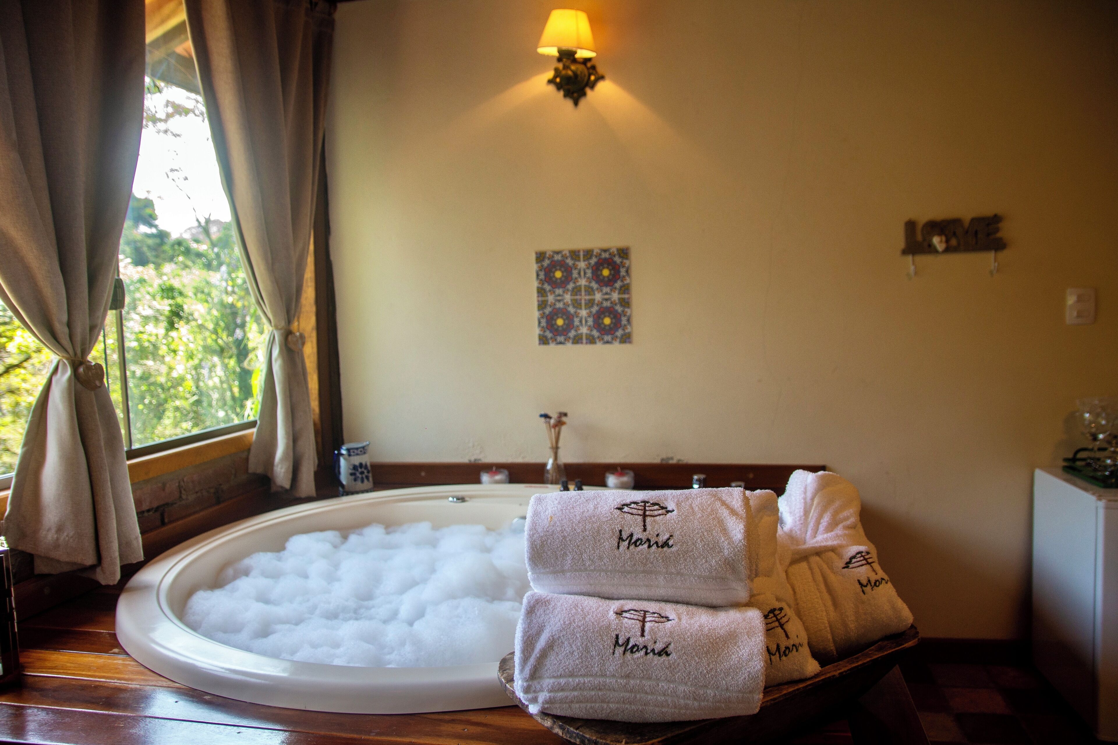 Ein Badezimmer mit einer Badewanne voller Bläschen, Handtüchern mit dem Namen "Maria" und einer Wandleuchte sowie einer kleinen Dekotafel.