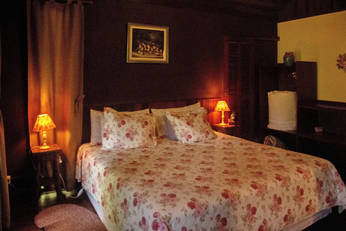 Quarto com colcha florida, duas luminárias de cabeceira, um quadro na parede e um banquinho próximo à cama.