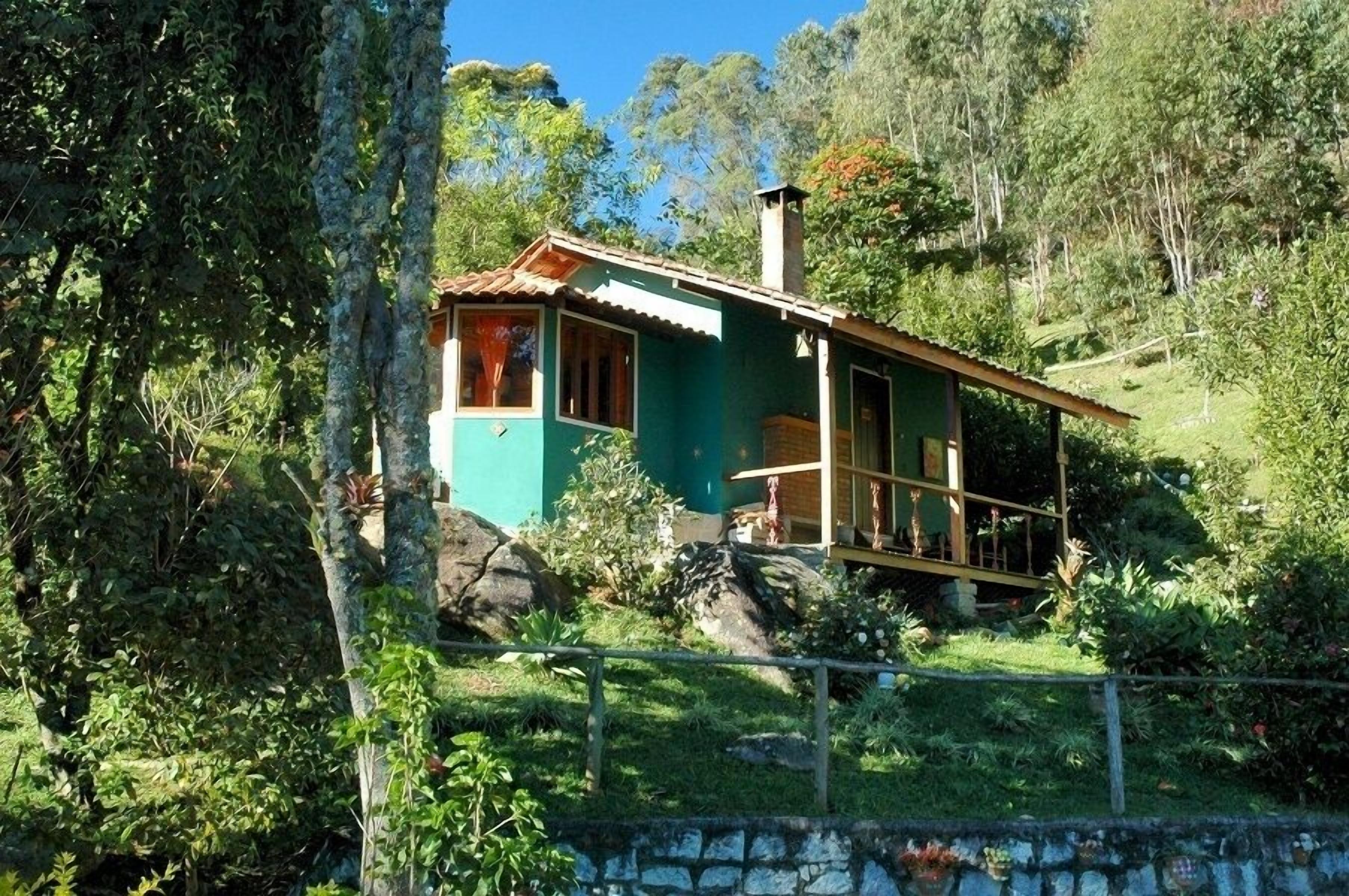 A small, single-story house with a green exterior and a red roof, surrounded by lush greenery and trees.