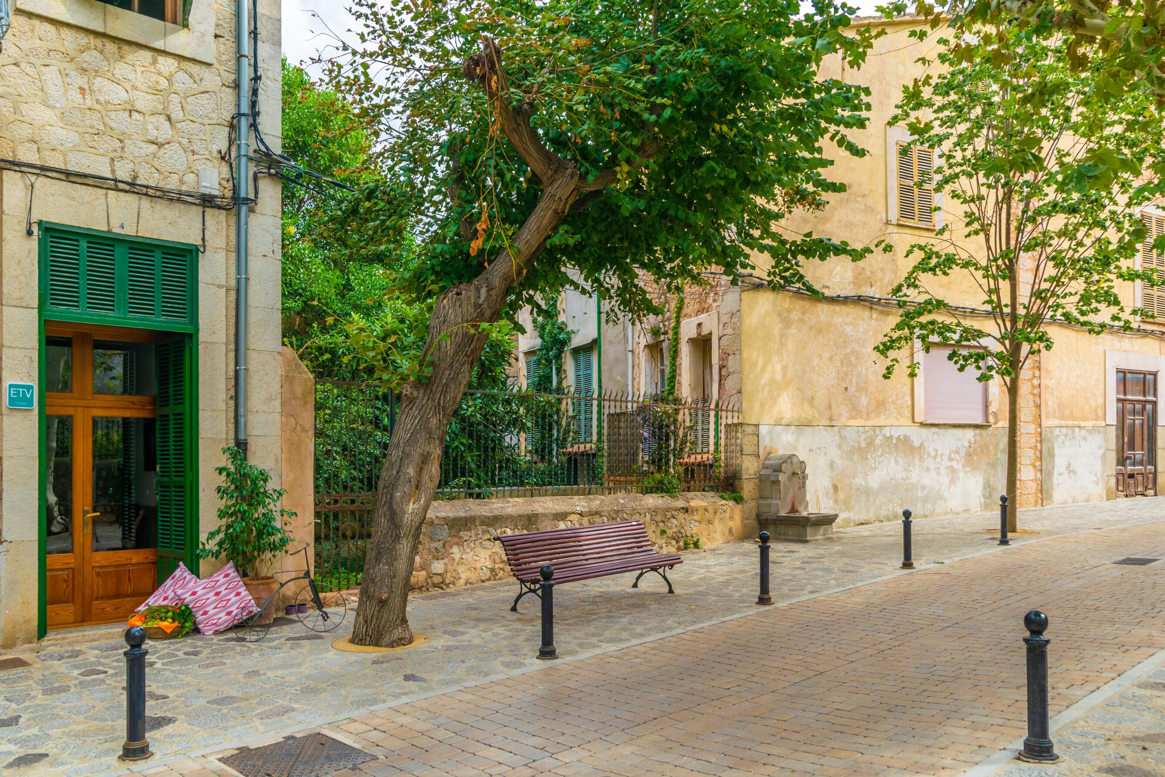 A cobblestone street with a bench, a tree, and a building with a stone facade and green shutters.