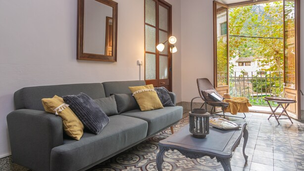 A living room with a grey sofa, a coffee table, and a chair. There is a window with a view of greenery and a building. - CA SA TIA (SÓLLER) - Townhouse with nice patio in the centre of Sóller. Free WiFi (Sóller)