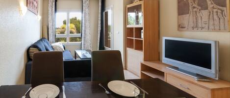 A dining area with a table set for two, a television on a stand, and a bookshelf.