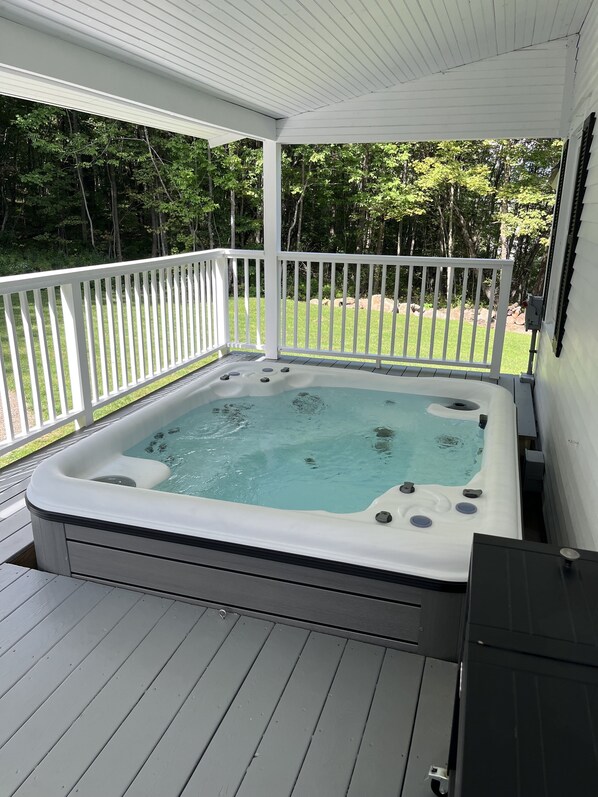 A hot tub on a wooden deck with a white railing, surrounded by a grassy yard and trees.