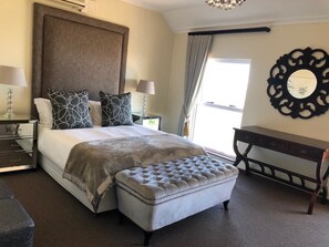 A neatly arranged bedroom with a large bed, two bedside tables with lamps, a tufted ottoman, a decorative mirror, and a view of the outdoors. - Cape Grandeur (Cape Town)