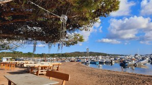 Ein Café am Strand mit Holztischen und -stühlen, freier Aussicht auf Boote im Wasser und hügeliger Landschaft im Hintergrund.