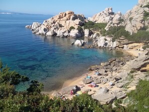 Una zona costera rocosa con agua cristalina, una playa de arena y gente relajándose.