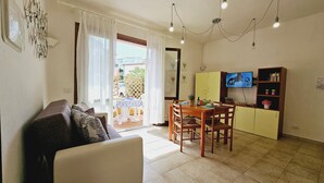 Ein Wohnzimmer mit einer Couch, einem Esstisch mit Stühlen, einer Küchenzeile mit einem gelben Schrank und einem Fernseher, sowie ein Balkon mit Außenblick.