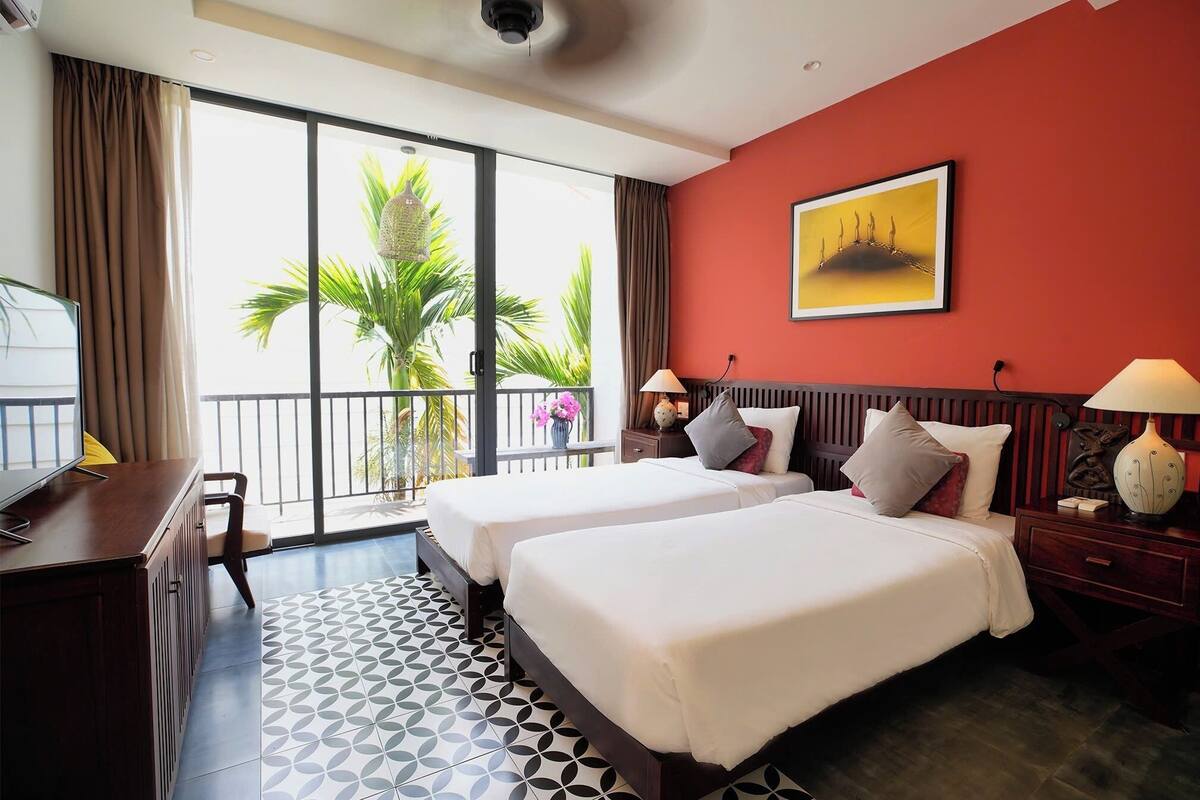 A hotel room with two beds, a desk, a chair, a TV, a balcony with a view of palm trees, and a red wall with a framed picture.