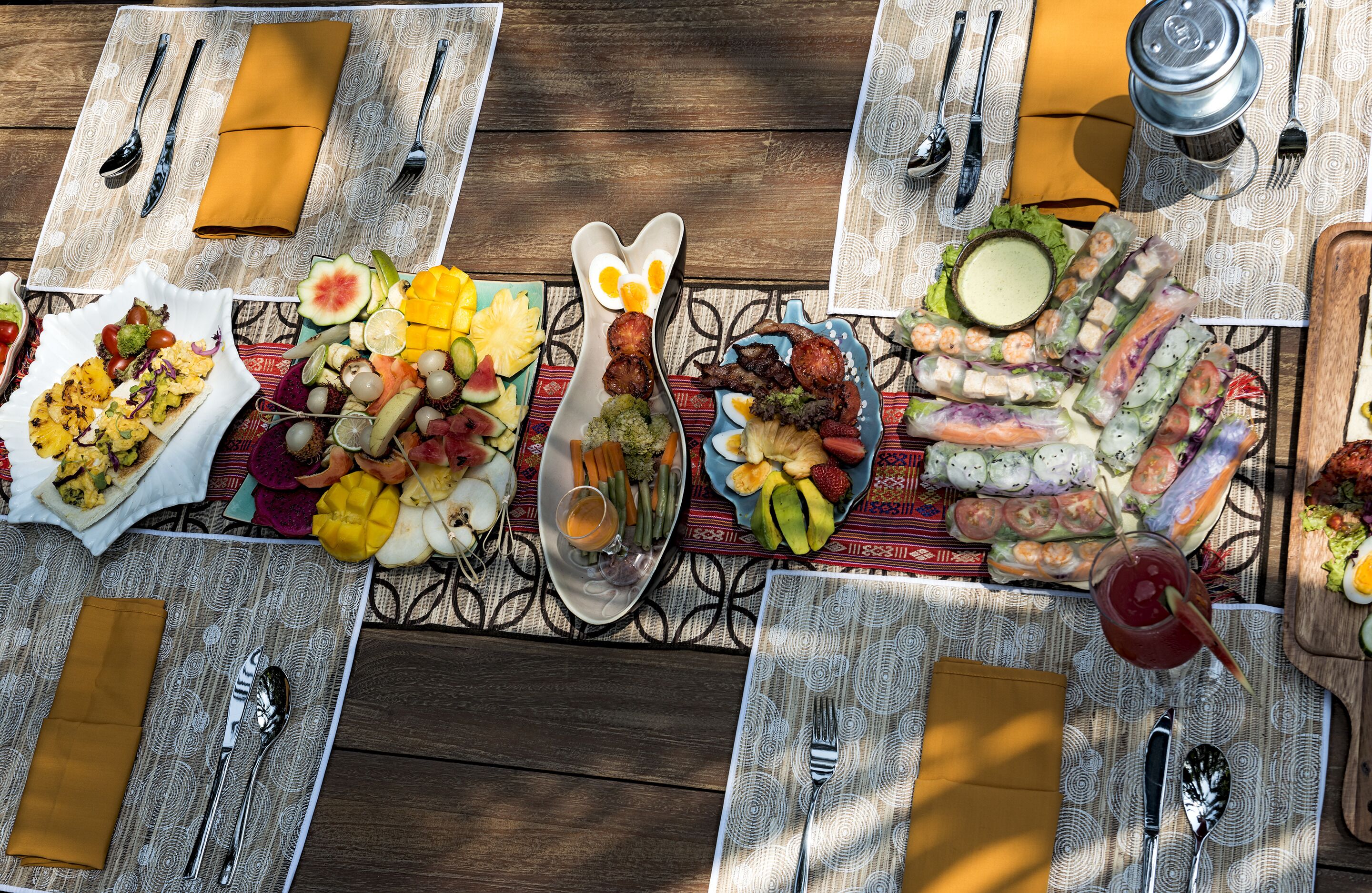 A table set with various dishes, including a plate of grilled vegetables, a bowl of fruit, and a plate of spring rolls.