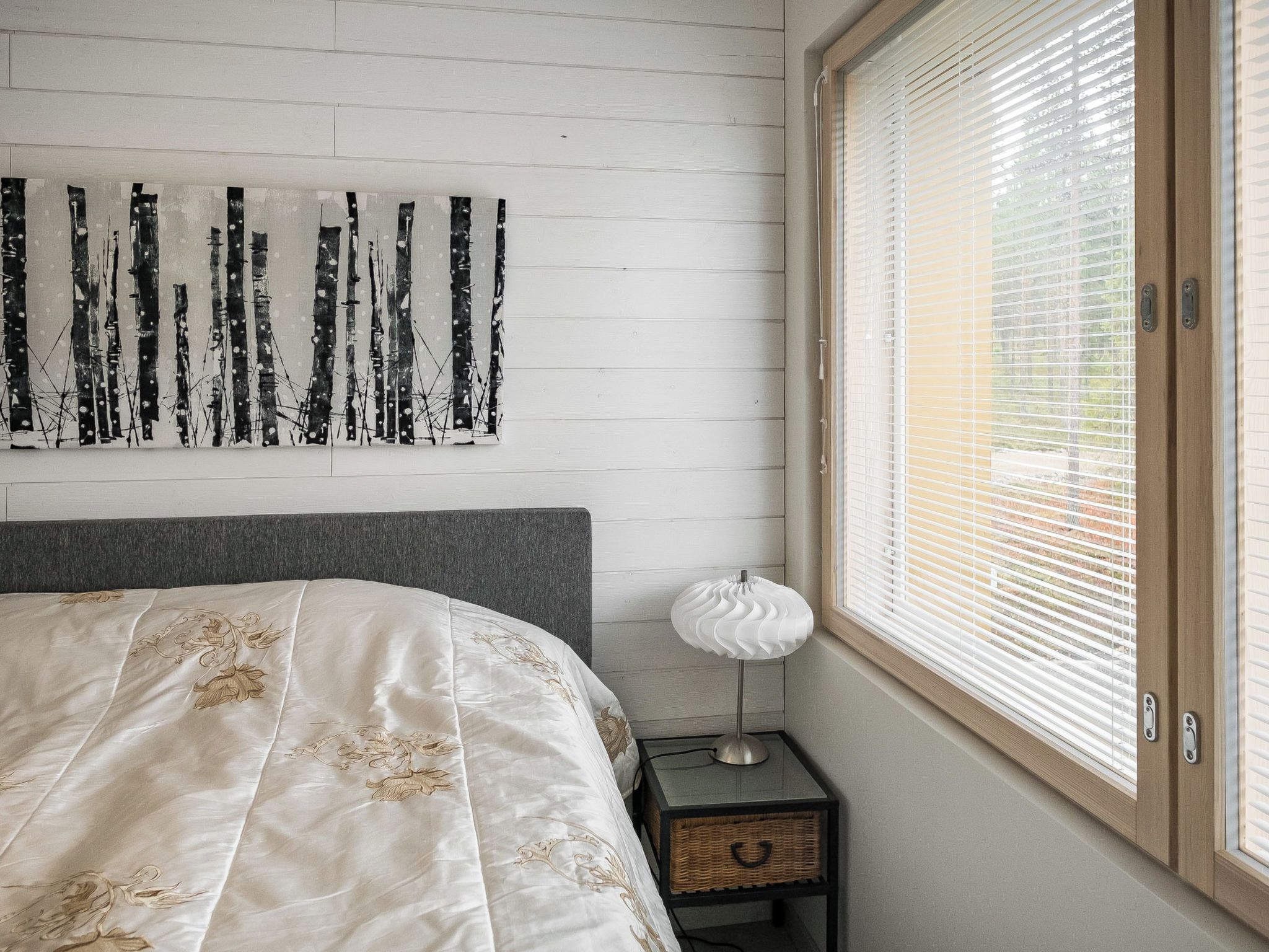 A bedroom with a bed, a wall-mounted abstract painting, a bedside lamp, and a window with blinds.
