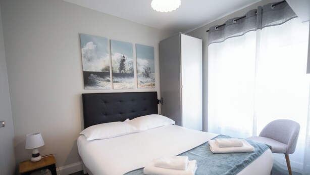 Un lit bien fait, recouvert d’une couverture bleue, une lampe de chevet et une chaise près de la fenêtre.