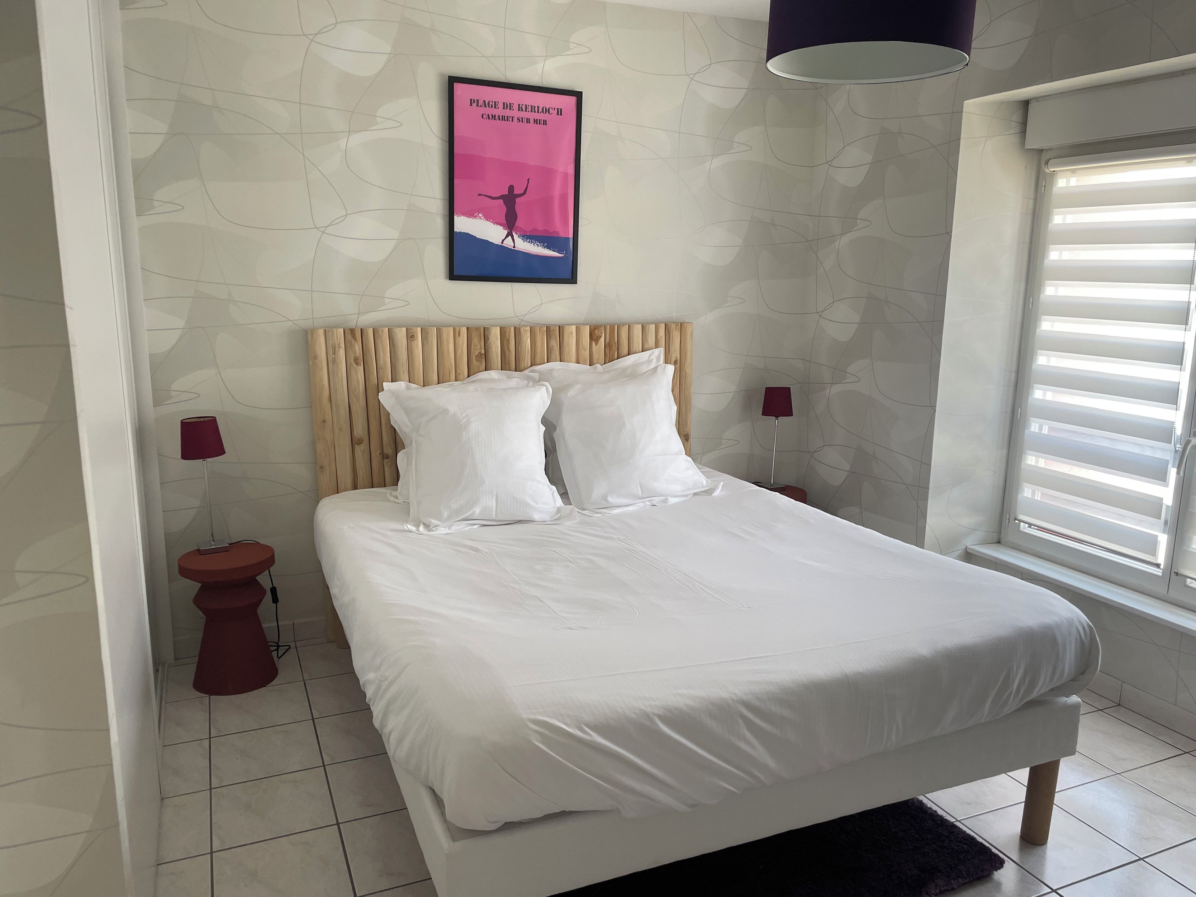 Quarto com cama, cabeceira de madeira, duas mesas de cabeceira, um criado-mudo, janela com persianas e um cartaz fixado na parede.