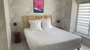 Quarto com cama, cabeceira de madeira, duas mesas de cabeceira, um criado-mudo, janela com persianas e um cartaz fixado na parede.